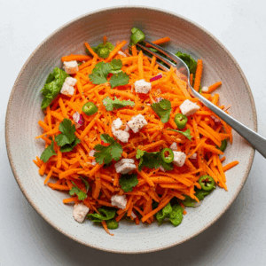 Bowl of crunchy carrot salad with shredded carrots, chopped green chili, fresh cilantro, leafy greens, cubes of cheese, and a fork on the side.