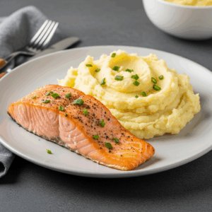 A plate of cooked salmon fillet seasoned with herbs and pepper, served alongside creamy mashed potatoes topped with chopped chives.