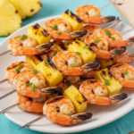 Close-up of grilled shrimp and pineapple skewers on a white plate with blue background, featuring juicy pink shrimp, charred golden pineapple chunks, green cilantro garnish, and skewers arranged neatly.