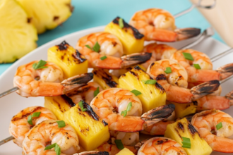 Close-up of grilled shrimp and pineapple skewers on a white plate with blue background, featuring juicy pink shrimp, charred golden pineapple chunks, green cilantro garnish, and skewers arranged neatly.